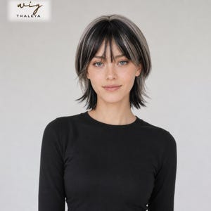 May include: A short, layered wig featuring a mix of black and silver-grey hair with wispy bangs. The hair is styled in a textured, shoulder-length cut. The text "wig THALEYA" is visible in the upper left corner of the image.