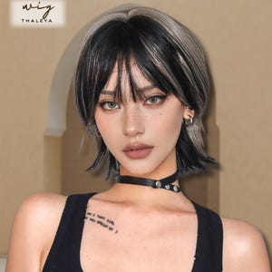 Puede incluir: Peluca corta y en capas con reflejos negros y plateados. La peluca presenta un flequillo recto y capas laterales. La marca "THALEYA" es visible. Se lleva un collarín negro.
