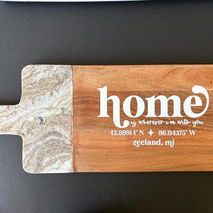 May include: Wooden serving board with a marble-effect handle and a rectangular wooden surface. The word "home" is written in white script, along with the phrase "is wherever I'm with you" and coordinates for Zeeland, MI.