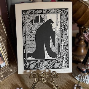 Peut inclure: Une estampe en noir et blanc d'une figure dans un jardin, exposée sur un chevalet doré. L'estampe présente des motifs floraux et foliacés complexes. Le texte "Irene Guinevere Made Her a Nun" est visible. Un chapelet de perles à motifs floraux est au premier plan.