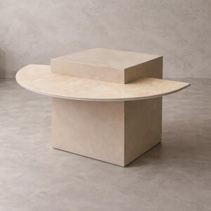 May include: A modern, beige coffee table with a geometric design. The table features a square base, a semi-circular top, and a smaller square element on top. The table's dimensions are approximately 30 inches in length, 30 inches in width, and 18 inches in height.