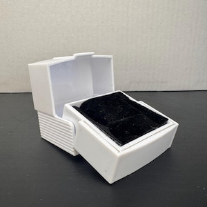 May include: A white plastic jewelry box with a hinged lid and a black velvet interior. The box is open, revealing the velvet-lined space for rings or other small items. The box has a rectangular shape with a textured side.