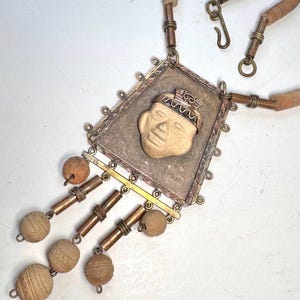 May include: A handcrafted necklace featuring a rectangular pendant with a carved face design. The pendant is accented with copper and brass elements, and includes dangling, textured, spherical beads. The necklace has a leather cord.