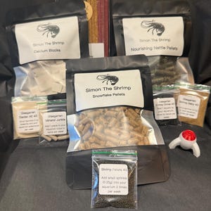 May include: A selection of shrimp food products in various clear and black packets. The image shows calcium blocks, nourishing nettle pellets, snowflake pellets, and other mineral and shrimp pellets. The brand name "Simon The Shrimp" is printed on the packaging.
