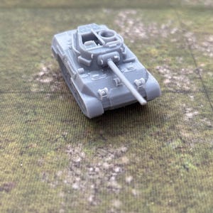 May include: A small, gray toy tank with a long barrel and detailed features. The miniature tank is set against a green and brown background, suggesting a battlefield or terrain.