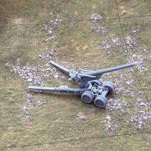 May include: A gray miniature artillery cannon with multiple wheels and a long barrel. The cannon is set against a backdrop of a green and brown terrain mat, suggesting a tabletop game or model.