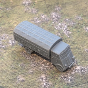 May include: A grey toy truck with a covered bed, set against a backdrop of a grassy terrain. The truck has a detailed front end and visible wheels, suggesting a miniature model or collectible item.