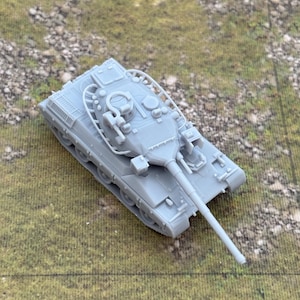 May include: A grey model tank with a long barrel and intricate detailing. The miniature tank is positioned on a textured green and brown surface, highlighting its design. The tank's tracks and turret are clearly visible.