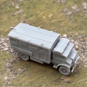 May include: A gray model truck with a boxy rear section and a detailed front cab. The truck has visible wheels, a front bumper, and a roof with ridges. The model is set on a textured, green and brown surface.