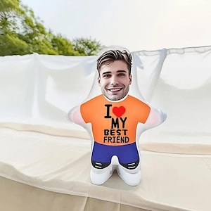 Puede incluir: Un cojín de peluche con forma personalizada que presenta una impresión fotográfica del rostro de una persona sonriente sobre un cuerpo estilo caricatura. El cuerpo viste una camiseta naranja con un corazón rojo y el texto negro I LOVE MY BEST FRIEND.