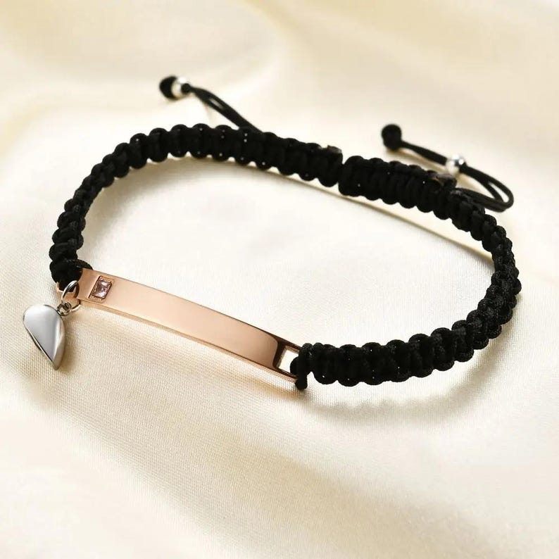 May include: A black braided bracelet with a rose gold rectangular plate and a silver heart charm. The plate has a small pink gem. The bracelet is on a white satin surface.