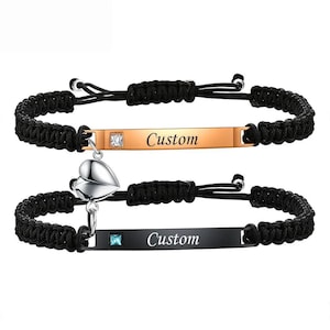 May include: Two matching black braided bracelets. One bracelet has a gold-colored rectangular plate with the word "Custom" and a small square gem. The other bracelet has a black rectangular plate with the word "Custom" and a small square blue gem. A silver heart charm connects the two bracelets.