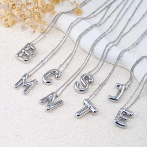Peut inclure: Colliers avec initiales argentées. Chaque collier présente un pendentif lettre différent, dont B, M, C, N, S, J, T et E. Les pendentifs sont reliés à de fines chaînes argentées. Les colliers sont présentés sur une surface blanche.