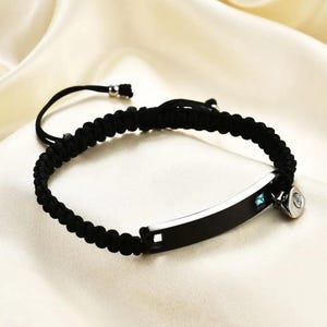 May include: Black braided bracelet with a silver-tone rectangular plate and a small, round charm. The plate has a small blue gem. The bracelet is adjustable and sits on a white satin surface.