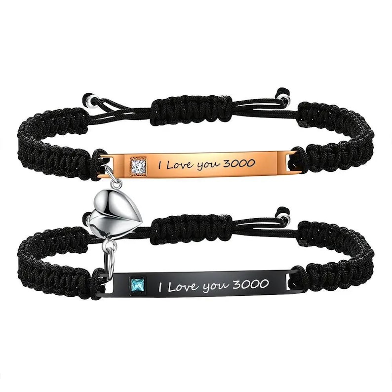 May include: Two matching black braided cord bracelets. One bracelet has a gold-colored rectangular bar with a clear square gem and the text "I Love you 3000." The other bracelet has a black rectangular bar with a blue square gem and the same text. A silver heart charm hangs from the second bracelet.