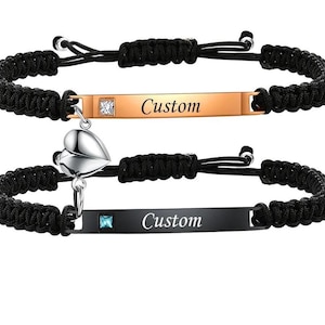 May include: Two matching black braided cord bracelets. One bracelet has a gold-colored rectangular plate with the word "Custom" and a clear square gem. The other bracelet has a black rectangular plate with the word "Custom" and a blue square gem, plus a silver heart charm.