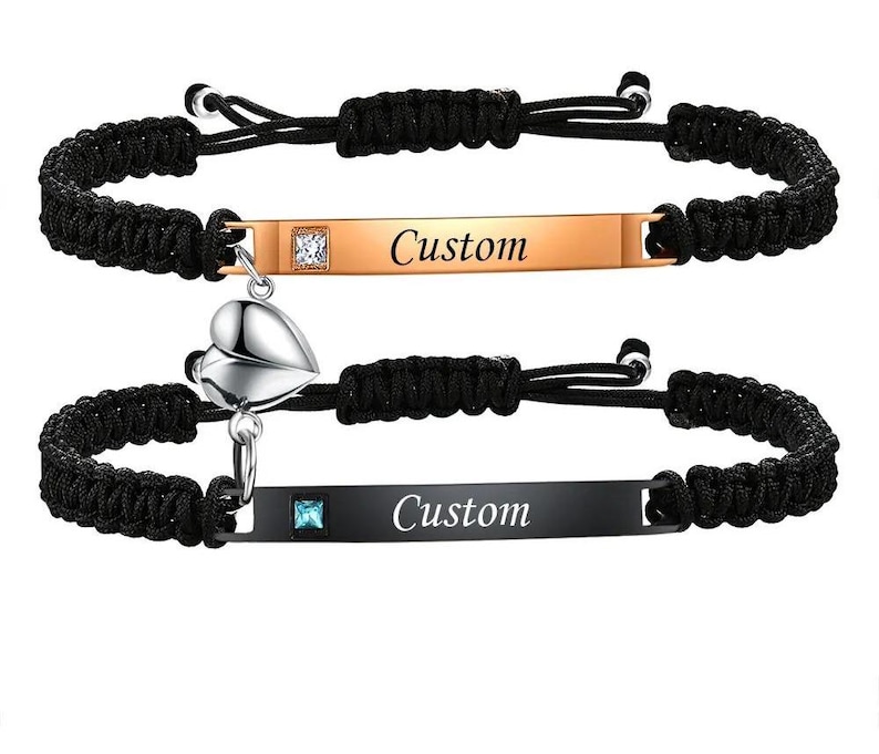 May include: Two matching black braided cord bracelets. One bracelet has a gold-colored rectangular plate with the word "Custom" and a clear square gem. The other bracelet has a black rectangular plate with the word "Custom" and a blue square gem, plus a silver heart charm.
