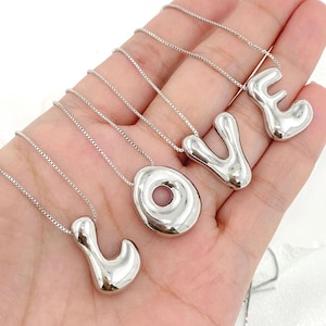 May include: Silver necklaces spelling "LOVE". Each letter is a separate pendant with a rounded, abstract design. The pendants hang from delicate silver chains, creating a modern jewellery set. The design is contemporary and eye-catching.