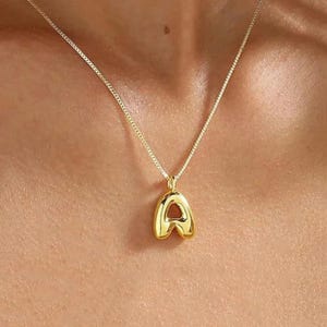 May include: A gold-coloured necklace featuring a pendant in the shape of the letter "A". The pendant has a rounded, bubble-like design with a central cutout. The chain is a fine box-style chain. The necklace is shown against a skin-toned background.