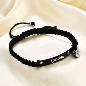 May include: Black braided bracelet with a silver-tone plate engraved with the word "Custom". The bracelet features a small blue gemstone and a decorative charm. The adjustable cord allows for a comfortable fit.