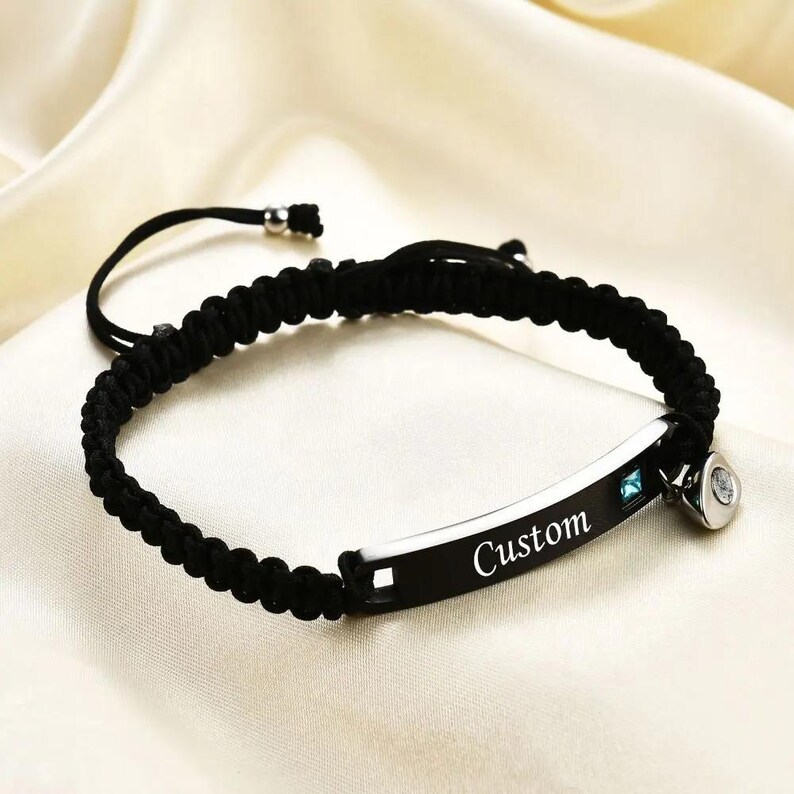 May include: Black braided bracelet with a silver-tone plate engraved with the word "Custom". The bracelet features a small blue gemstone and a decorative charm. The adjustable cord allows for a comfortable fit.