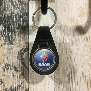 May include: Black leather keychain with a silver ring and a circular emblem. The emblem features a red griffin with a crown on a blue background, and the word "SAAB" in white.