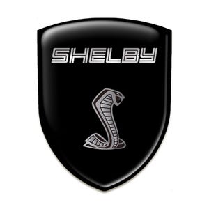 May include: A black shield-shaped emblem featuring the word "SHELBY" in silver. Below the text is a silver cobra snake emblem. The emblem has a glossy finish and a white background.