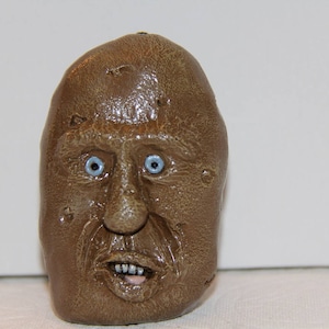 Könnte beinhalten: Eine braune Keramik-Gesichtsskulptur mit blauen Augen und offenem Mund. Das Gesicht hat eine strukturierte Oberfläche und ist ein einzigartiges Kunstwerk. Die Skulptur ist ein skurriles und ungewöhnliches Dekorationsstück.