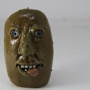 Puede incluir: Una escultura de cerámica marrón de una patata con cara humana. La patata tiene ojos azules, una boca ancha y la lengua fuera.