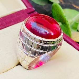 May include: A silver ring featuring a large, smooth, oval-shaped cabochon ruby gemstone. The metal band has a distinct grid-like pattern of recessed lines carved into the surface, creating a series of small, raised square sections around the stone.