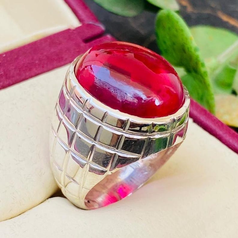May include: A silver ring featuring a large, smooth, oval-shaped cabochon ruby gemstone. The metal band has a distinct grid-like pattern of recessed lines carved into the surface, creating a series of small, raised square sections around the stone.