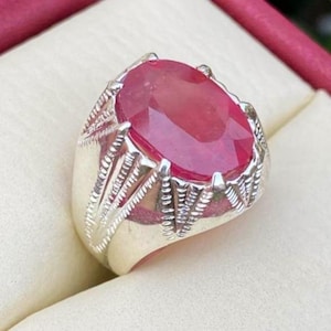 May include: A silver ring featuring a large, oval-cut ruby gemstone. The ruby is a deep red color and is set in a detailed silver band with intricate designs. The ring is displayed on a cream-colored surface.