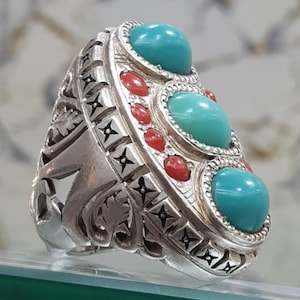 May include: A silver ring with an ornate design. The ring features three large, oval turquoise stones and smaller red stones. The band has intricate carvings and geometric patterns. A unique piece of jewelry.