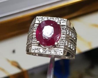 May include: A silver ring featuring a large, oval-cut ruby. The ring is embellished with smaller, clear gemstones around the ruby and along the band. The ring is held by tweezers, showcasing its intricate design and the deep red hue of the ruby.