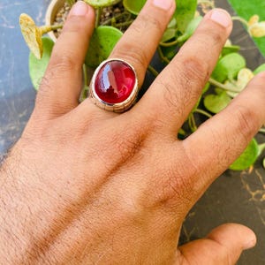 May include: A silver ring featuring a large, oval-shaped, deep red cabochon gemstone. The metal band has a textured, grooved design along the sides, and the stone is held in place by a smooth, polished silver bezel setting that encircles the gem.