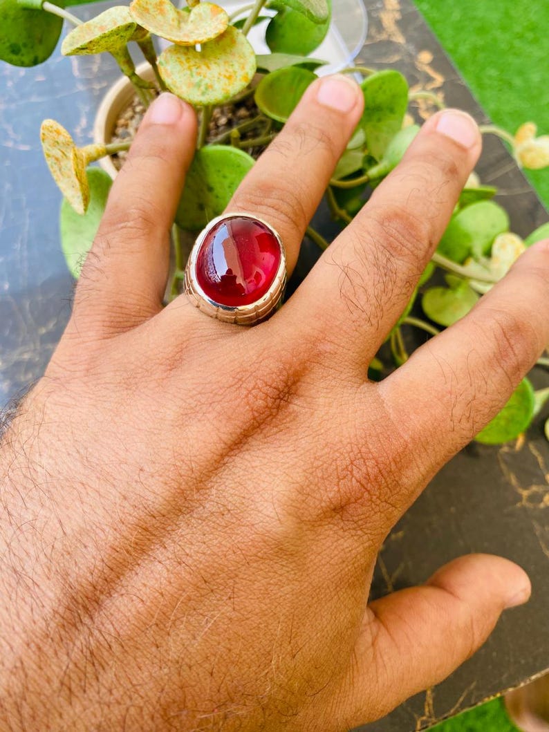 May include: A silver ring featuring a large, oval-shaped, deep red cabochon gemstone. The metal band has a textured, grooved design along the sides, and the stone is held in place by a smooth, polished silver bezel setting that encircles the gem.