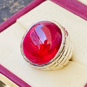 May include: A silver-toned ring featuring a large, smooth, oval-shaped cabochon gemstone in a deep, vibrant red color. The metal band displays a textured, grid-like pattern along its sides, providing a structured contrast to the polished stone.