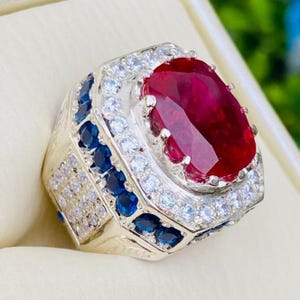 May include: A silver-toned ring featuring a large, oval-cut deep red gemstone at the center. The stone is surrounded by a halo of small, clear white stones, with additional rows of deep blue stones and clear white stones set along the band.