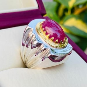 May include: A silver ring featuring a large, oval-shaped, cabochon-cut magenta ruby gemstone set in a gold-toned bezel. The band of the ring has a wide, polished silver surface with vertical grooved detailing along the sides of the setting.