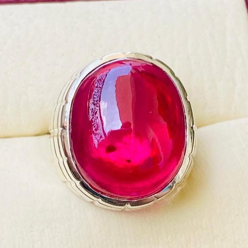 May include: A silver-toned ring featuring a large, oval-shaped, smooth cabochon-cut vibrant pink gemstone. The metal band displays a segmented, notched design around the perimeter of the setting, framing the polished, translucent pink stone.