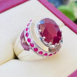 Puede incluir: Un anillo de tono plateado con una gran piedra preciosa de color rojo intenso y corte ovalado en el centro. El engaste está rodeado por un halo de piedras rojas más pequeñas de corte redondo incrustadas en la banda, creando un contraste vibrante.