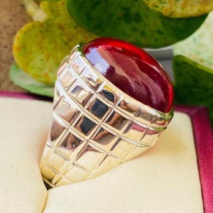 May include: A silver-toned ring featuring a large, smooth, deep red cabochon gemstone set into a wide band. The metal band is decorated with a prominent grid-like, waffle-patterned texture that extends from the base up to the stone setting.