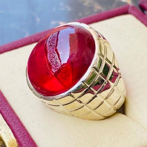May include: A gold ring featuring a large, smooth, cabochon-cut deep red gemstone set in a band with a textured, grid-like pattern. The metal band has a polished finish, and the vibrant red stone displays a glossy surface with internal reflections.