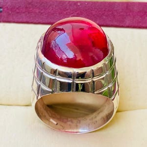 May include: A silver-toned ring featuring a large, smooth, cabochon-cut deep red gemstone set in a wide band. The metal band displays a structured, grid-like pattern of recessed lines around the setting, providing a textured appearance.