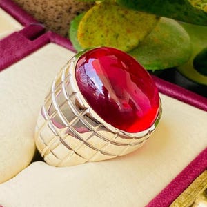 May include: A silver-toned ring featuring a large, smooth, oval-shaped cabochon red gemstone set in a wide band. The metal band displays a textured, grid-like pattern of recessed squares across its surface, providing a geometric contrast to the stone.
