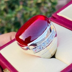 Könnte beinhalten: Ein silberfarbener Ring mit einem großen, glatten, ovalen, tiefroten Cabochon-Stein, der oben prominent eingefasst ist. Das Band ist mit einer Reihe kleiner, klarer, funkelnder Steine verziert, die in die Seite der Metallfassung eingelassen sind.