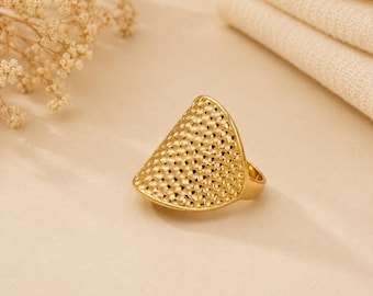 May include: A gold-toned ring featuring a wide, triangular-shaped face with a textured, dimpled surface. The metal has a polished finish, and the band is integrated into the back of the wide, curved, and slightly pointed front design element.