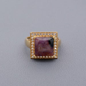 May include: A gold ring featuring a square-cut, purplish-red gemstone with a black inclusion. The gemstone is surrounded by a square frame of small, clear stones. The ring has a simple, rounded band.