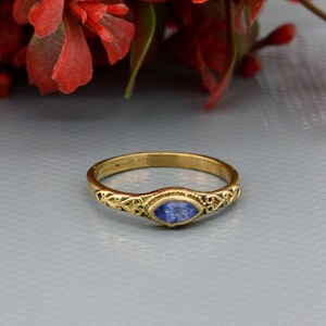Könnte beinhalten: Ein goldener Ring mit einem blauen, ovalen Edelstein. Der Ring hat ein verziertes Design am Band und ist vor einem hellgrauen Hintergrund platziert. Rote Blumen sind im Hintergrund zu sehen.