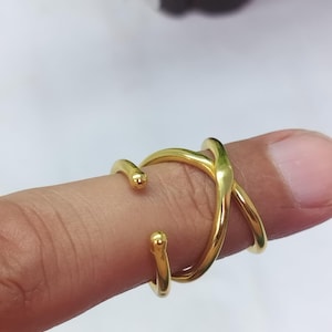Puede incluir: Un anillo abierto de tono dorado con un diseño cruzado y dos extremos lisos y redondeados. La banda es pulida y reflectante, creando un bucle continuo que se superpone a sí mismo, formando una figura en forma de X sobre el dedo.